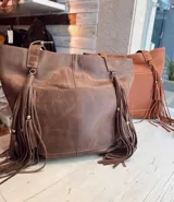 Cartera tipo tote bag de cuero marrón con diseño de flecos laterales largos, bolsillo frontal externo y costuras visibles.