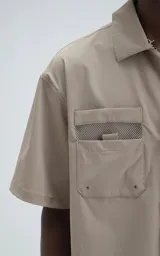 Camisa de manga corta color beige, estilo técnico, confeccionada en microfibra liviana. Cuenta con doble bolsillo frontal, uno con detalle de malla y otro con cierre, y broches en lugar de botones.