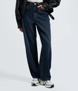 Pantalón de jean corte baggy con cintura media, diseño de cinco bolsillos y cierre frontal con cremallera y botón. Presenta un lavado oscuro sobretinta y una trabilla con colgante metálico desmontable.
