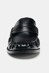 Mocasín plano negro de cuero liso, con antifaz, copete subido y detalle de fruncidos en la parte delantera. Punta redonda y suela de 2,5 cm de altura.
