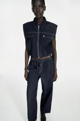 Chaleco de denim azul oscuro con cuello solapa, cierre frontal con cremallera y bajo elástico abullonado. Presenta tapetas delanteras y costuras en contraste.