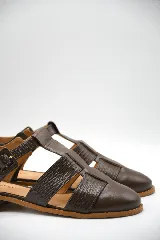 Zapato tipo cangrejera de cuero color marrón, con diseño de tiras entrelazadas, punta cerrada y pulsera con hebilla ajustable al tobillo. Cuenta con un taco bajo de madera.