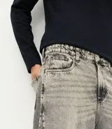 Pantalón jean de corte super baggy con piernas amplias, confeccionado en denim de algodón. Presenta un acabado lavado grisáceo, ruedo desflecado y detalle de costura frontal tipo nesga.