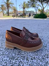 Mocasines de cuero marrón con plataforma, detalle de tachas metálicas y textura granulada en el talón y la puntera.