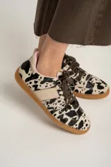 Zapatillas bajas de cuero con estampado de vaca en blanco y negro, cordones marrones y suela de goma color caramelo.