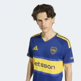 Camiseta de futbol Adidas de la primera equipacion de Boca Juniors, de color azul con la caracteristica franja horizontal amarilla en el pecho. Presenta cuello en V, mangas cortas con las tres tiras de Adidas en los hombros y el logo de la marca y el escudo del club bordados en el frente.