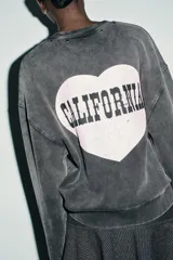Sudadera de cuello redondo y manga larga, color gris oscuro con efecto lavado. Presenta aplicación de tachas metálicas alrededor del cuello y estampado frontal con el texto "CALIFORNIA USA" en blanco.