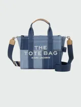 Cartera tipo tote bag de Marc Jacobs, confeccionada en denim con diseño de patchwork en diferentes tonos de azul. Presenta el logo estampado en el frente, doble asa superior y correa de hombro ajustable y extraíble en color azul marino con detalles en cuero tostado.