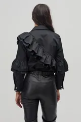 Camisa negra de popelina de algodón con cuello clásico y cierre frontal de botones de nácar. Presenta volados dobles que recorren el frente y la espalda desde el hombro hasta la cintura, y volados adicionales en las mangas a la altura de los codos.