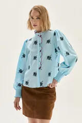 Blusa de algodón en color celeste con estampado floral en negro. Presenta cuello alto con volados y borde en color rojo, mangas abullonadas con puños elásticos tipo smock y cierre frontal con botones.