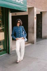 Chaqueta de denim azul con cuello redondo, manga larga y cierre frontal con botones dorados. Presenta costuras verticales decorativas y puños con botones a juego.