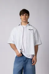 Camisa de manga corta a rayas beige y blancas, de corte boxy y liviana, con cuello camisero, cierre frontal de botones y pequeño texto bordado "BUILD IT BOLD" en el pecho.