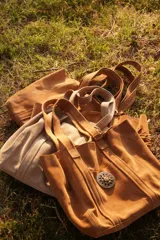 Cartera tipo tote de gamuza color marrón, con diseño amplio y relajado. Presenta un bolsillo frontal externo con cierre de broche metálico y una hebilla decorativa circular, además de un detalle de borla colgante en el lateral.