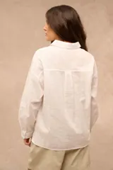 Camisa blanca de manga larga con cuello camisero, cierre frontal de botones y un bolsillo aplicado en el pecho.