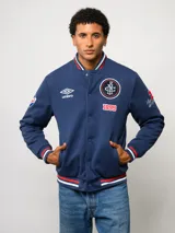 Campera bomber azul marino estilo retro, con cierre frontal de botones a presión, cuello, puños y ruedo con tejido acanalado tricolor. Presenta el logo de Umbro y el escudo del Club Nacional de Football bordados en el pecho, junto con el año 1899 y parches decorativos en las mangas.