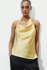 Top de cuello halter con escote fluido y espalda descubierta. Presenta un diseño satinado en color amarillo con estampado de lunares pequeños y un aplique metálico decorativo en el cuello.