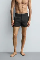 Bañador de hombre color negro, de largo medio, confeccionado en tejido técnico. Presenta cintura elástica ajustable con cordón interior, trabillas laterales, bolsillos frontales y bolsillo trasero con cierre de cremallera.