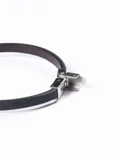 Pulsera de cuero negro con apliques de acero inoxidable plateado. En el centro presenta un dije rectangular con un águila grabada.