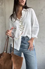 Camisa de lino color blanco, de corte holgado y manga larga con puños arremangados. Presenta cuello clásico, cierre frontal con botones y detalle de botones decorativos en el lateral inferior.