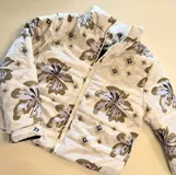 Campera puffer color beige con estampado floral y de logotipos en tonos marrones y grises, cuello alto y cierre frontal con cremallera.