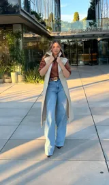 Trench coat de color beige, con diseño de doble botonadura, solapas amplias y cinturón ajustable en la cintura. Cuenta con mangas largas con trabillas en los puños y bolsillos laterales.