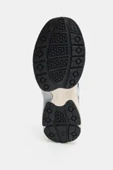 Zapatilla deportiva estilo running con combinación de materiales y colores. Presenta paneles en azul marino, plateado y detalles en amarillo neón, con suela de plataforma en tono beige y tirador en el talón.