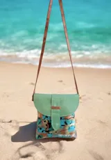 Cartera y bandolera de cuero Emi línea Ottenta, diseñada por Carolina de Cunto con arte pintado a mano. Presenta una solapa semicircular de cuero verde con textura símil serpiente y broche imán, cuerpo principal con estampado artístico multicolor en tonos celestes, marrones y negros, y correa larga marrón fina.