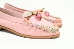 Mocasín plano de cuero color rosado, con punta redondeada y taco bajo. Presenta un detalle artesanal de tiras entrelazadas en tonos nude, verde y rosado alrededor del empeine, rematado con un aplique colgante central con piedra transparente.