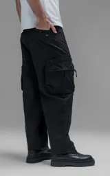 Pantalón cargo oversize de corte relajado, confeccionado en textil técnico resistente de color negro. Presenta bolsillos laterales cargo de gran tamaño con cierres metálicos, bolsillos laterales superiores y bolsillos traseros con broches a presión. Incluye cinturón integrado de cinta de seguridad con hebilla de encastre técnica.