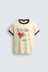 Camiseta de corte regular, color crema, con cuello redondo y ribetes en las mangas y cuello en color negro. Presenta un estampado frontal con un corazón rojo y texto en negro que dice "Everything beats for something".