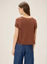 Remera tejida color beige de corte relajado, con mangas cortas y detalle de costuras frontales marcadas. Incluye un pequeño bordado distintivo en el borde inferior.