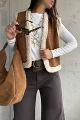 Chaleco de gamuza color camel con interior de corderito sintético, diseño sin mangas, borde frontal con ribete de corderito y detalle de tachas metálicas decorativas.