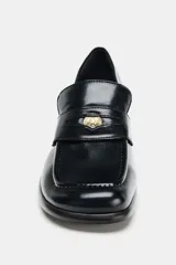 Mocasín de mujer en color negro con acabado liso, diseño de punta cuadrada y tacón en bloque. Presenta un detalle metálico decorativo en el antifaz frontal.