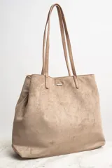 Bolso tipo tote de gran tamaño en color camel, confeccionado en cuero vegano con acabado efecto gamuza. Presenta doble asa larga para llevar al hombro y una placa metálica pequeña con el logo de la marca en el frente.