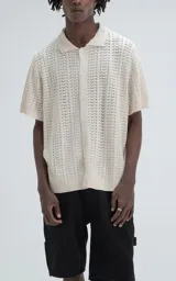 Camisa de tejido de punto color beige, manga corta, con cuello polo y cierre frontal de botones. Presenta un calce regular y un patrón calado en zigzag.