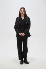 Trinchera de corte corto y diseño clásico, confeccionada en poliéster color negro. Presenta solapas amplias, cierre frontal cruzado con lazo ajustable en la cintura, charreteras en los hombros y puños con trabillas.