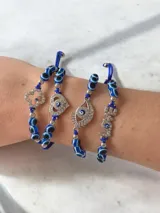 Pulsera de hilo azul con cuentas de ojos turcos y dijes metálicos con strass, incluyendo formas de corazón, ojo y la palabra love. Cuenta con un sistema de cierre ajustable mediante nudo corredizo.