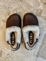 Zueco de cuero color suela con interior forrado en piel sintética blanca. Presenta una tira trasera ajustable con hebilla metálica y suela de goma negra con relieve.