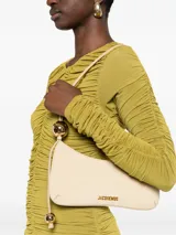 Bolso tipo baguette Jacquemus modelo Le Bisou Perle, de cuero con efecto cocodrilo en color beige. Presenta una correa delgada ajustable con dos esferas metálicas doradas decorativas y el logotipo de la marca en letras doradas en el frente.