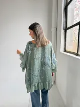 Blusa verde de lino con estampado floral en tonos azules y grises, escote redondo, mangas 3/4 con volados y ruedo asimétrico con volado fruncido.
