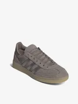 Championes Adidas modelo Handball Spezial de perfil bajo, confeccionados en gamuza color marrón topo con las clásicas tres tiras y detalles metalizados en tono plateado, inscripción lateral en relieve y suela de caucho translúcido.