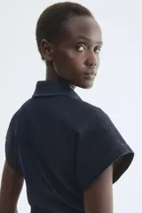 Vestido midi de denim azul oscuro, con cuello camisero, escote en V y cierre frontal de botones metálicos. Cuenta con mangas corta y frunces laterales en la cintura para un ajuste entallado.