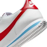 Championes Nike modelo Cortez, de diseño clásico en color blanco con el icónico swoosh lateral en rojo. Presenta detalles en rojo en el talón y una franja azul en la entresuela, con suela dentada característica.