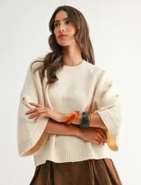 Buzo de punto beige con mangas tres cuartos anchas, cuello redondo y detalle de botones en las mangas.