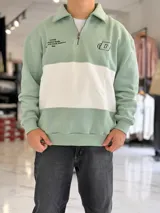Buzo de felpa verde menta y blanco con cuello polo, cierre frontal parcial, manga larga y puños elásticos. Presenta detalles estampados y bordados en el pecho.