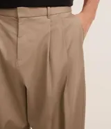 Pantalón de vestir color beige, de corte amplio estilo japonés, con cintura con trabillas, cierre tradicional, pliegues frontales, bolsillos laterales y traseros, y efecto satinado.