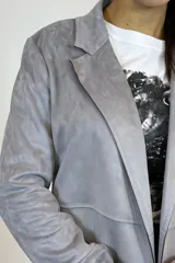 Chaqueta de corte recto en color gris, confeccionada en material tipo gamuza sintética. Presenta solapas clásicas, cierre frontal, mangas largas y dos bolsillos frontales aplicados.