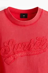 Remera de algodón en punto pesado, color rojo con efecto lavado, cuello redondo acanalado, hombros caídos y estampa frontal con relieve que dice 'Sunset'.