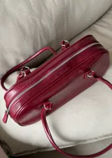 Cartera de mano tipo bowling color bordó, confeccionada en material sintético con textura arrugada. Cuenta con doble asa fija y cierre superior.