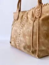 Cartera tipo tote de cuero color camel, con diseño artesanal que presenta costuras decorativas visibles en los laterales y el frente. Cuenta con doble asa de hombro y cierre superior.